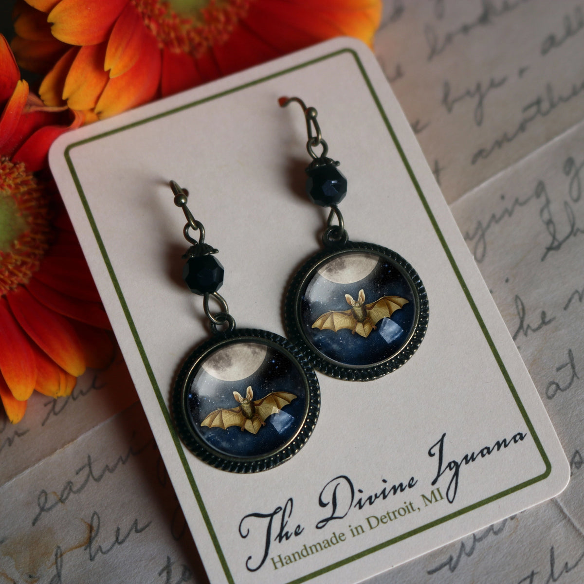 A pair of gothic-style bat drop earrings displayed on a Divine Iguana branded card. The earrings feature dark metal hooks, dark beads, and round bezel-set dangles showing a golden bat flying against a dark blue night sky with a full moon on Halloween.