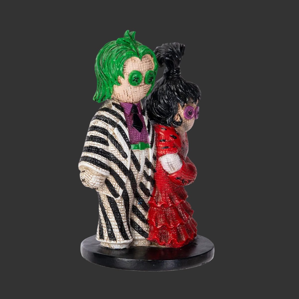 A small Pinhead Monsterfigurine of two characters, Beetlejuice and Lydia standing together on a round black base against a plain white background. One figure has bright green hair, a black-and-white striped jacket, and a purple shirt; the other has black hair, a red dress, and purple glasses.