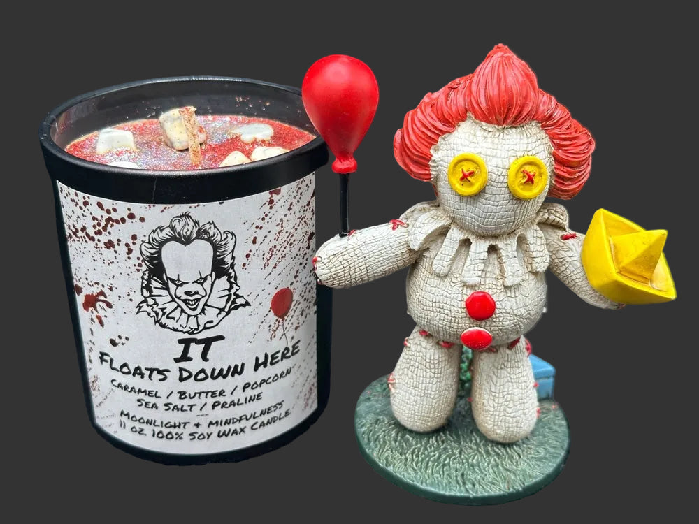 A Halloween-themed Pennywise candle and figurine set. The candle in a black jar has a white label with Pennywise the Clown illustration and the text 'IT FLOATS DOWN HERE'.