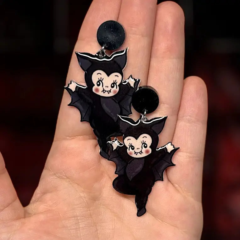 A pair of black vampire-shaped earrings with cute kewpie cartoon faces, held in a person's hand against a dark blurred background.