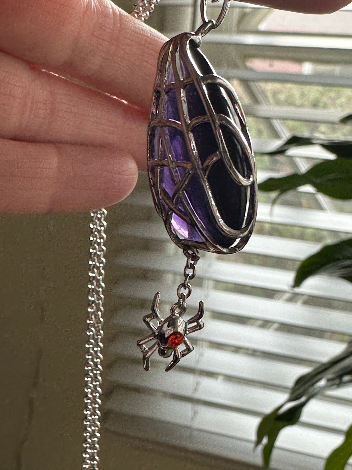 A close-up of a hand holding an Anne Stokes gothic-style pendant necklace. The pendant features a silver filigree cage enclosing a purple and black marbled stone, with a dangling silver spider charm that has a small red gem on its back.