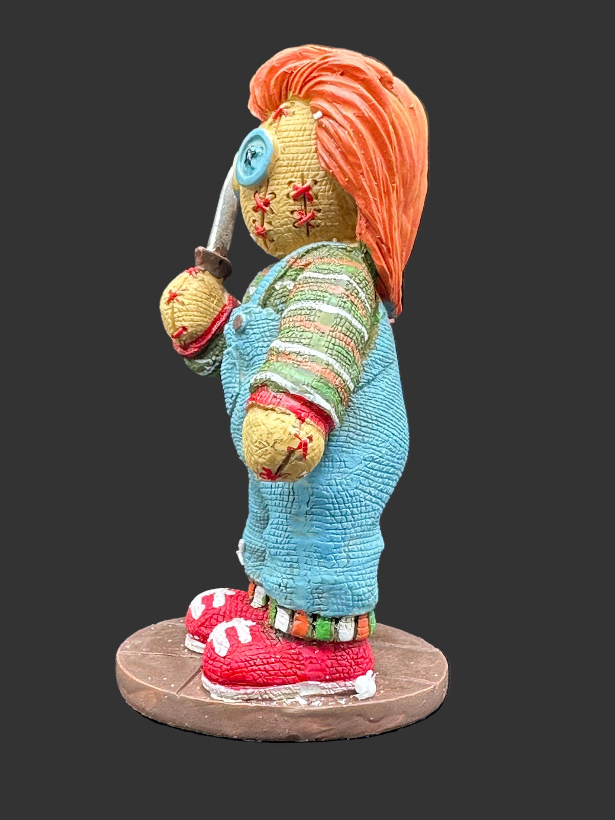 A horror-themed collectible figurine of a creepy doll with orange hair, a blue button eye, and a knife, wearing blue overalls and red sneakers on a circular base.