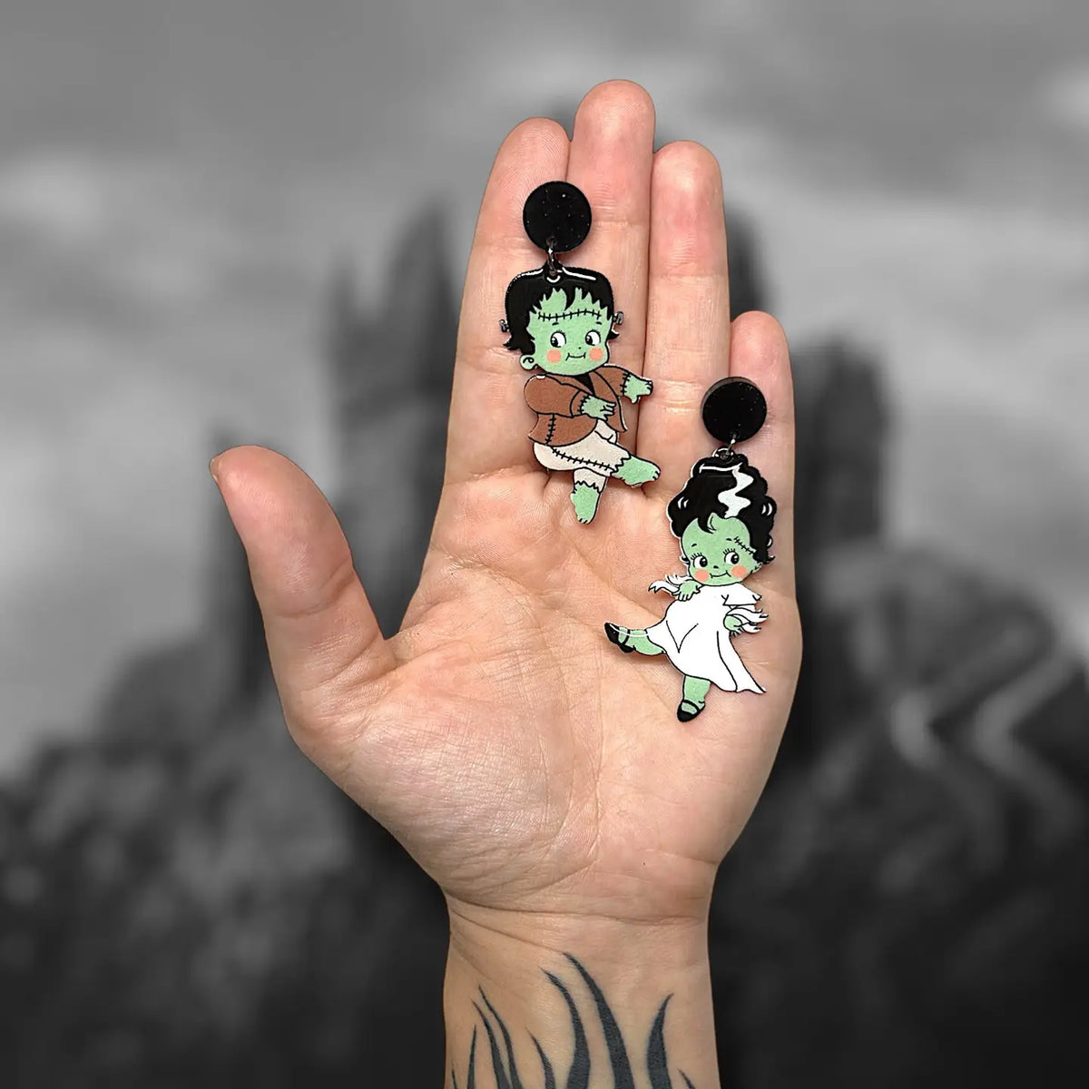 A hand holding two dangling earrings with black round studs and cartoon-style green-skinned character Frankenstein and his bride Elizabeth charms, against a blurred black-and-white background.