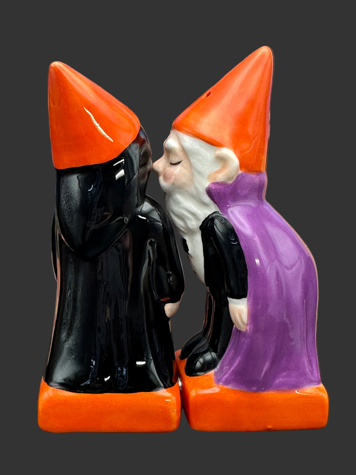 A pair of ceramic Halloween gnomes kissing, one in a black robe with an orange hat and base, and the other in a purple robe with an orange hat and base, set against a blurred Halloween village backdrop.