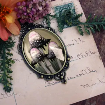 An oval gothic-style metal pendant or brooch with a decorative black filigree frame, featuring a fantasy illustration of three black mushrooms with pink and white spots on a vintage-style letter background with flowers and greenery.