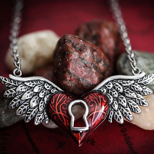 A gothic-style winged heart pendant necklace with a red enamel heart, silver-toned metal wings, and a lock-shaped cutout, displayed on red rocks over a dark red textured background.
