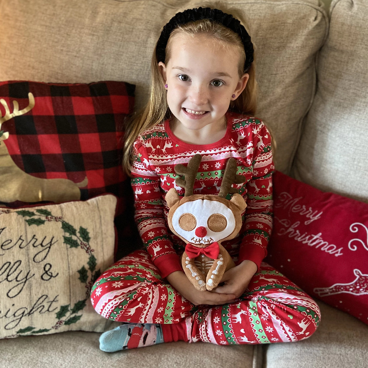 Child in festive pajamas holding a reindeer plush toy on a couch with Christmas decor.