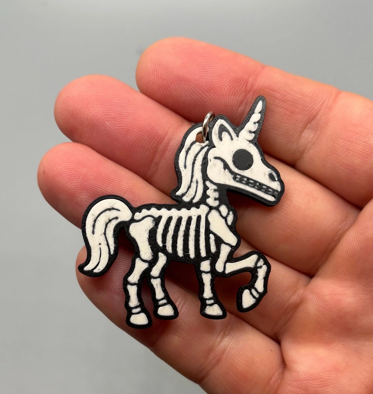 A small skeleton unicorn charm or keychain with black and white coloring, held in a person's hand against a plain light gray background.