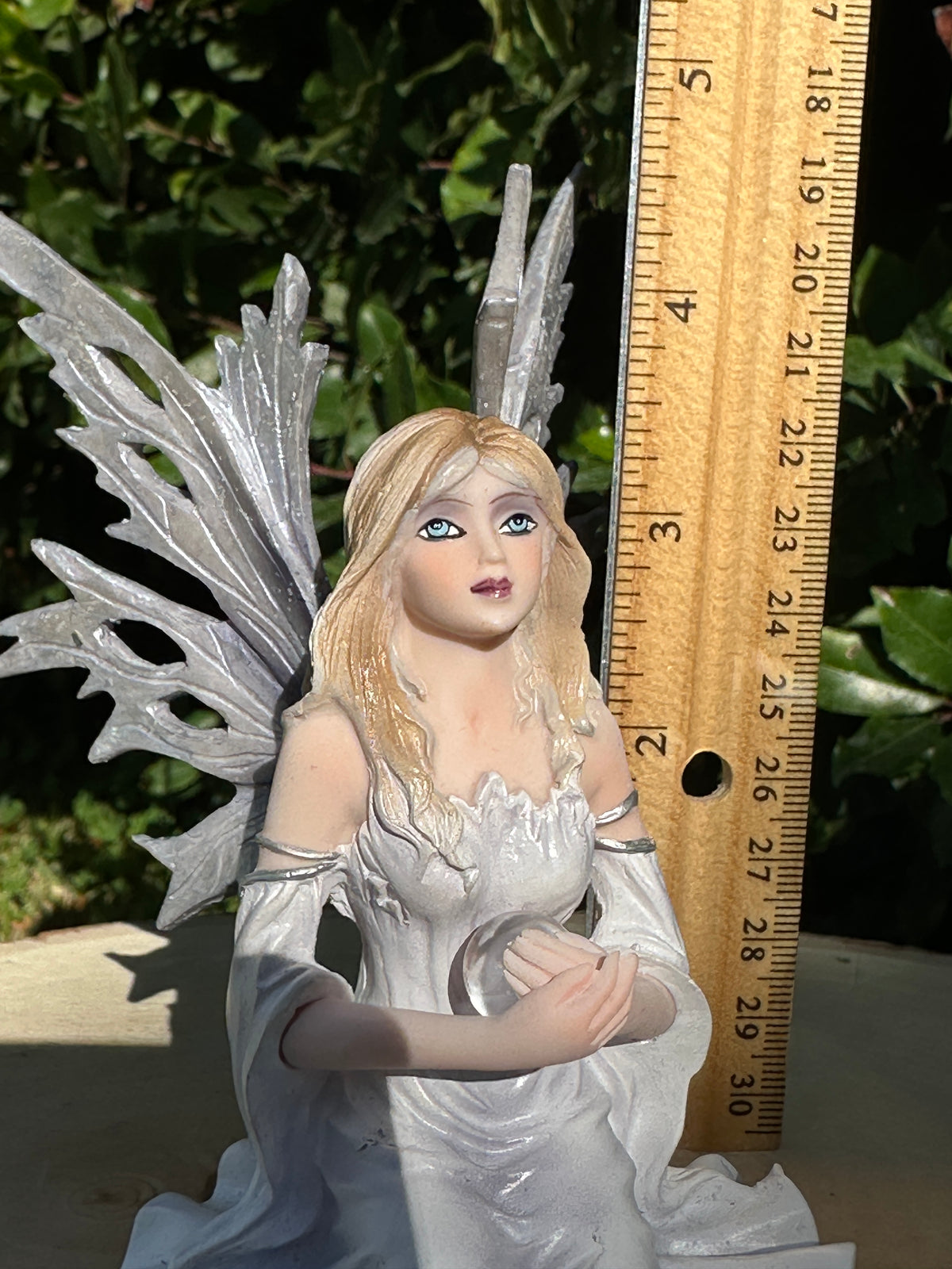 A small Dream Eden winter fairy figurine with silver wings and long blonde hair, shown next to a wooden ruler for scale outdoors with green foliage in the background.