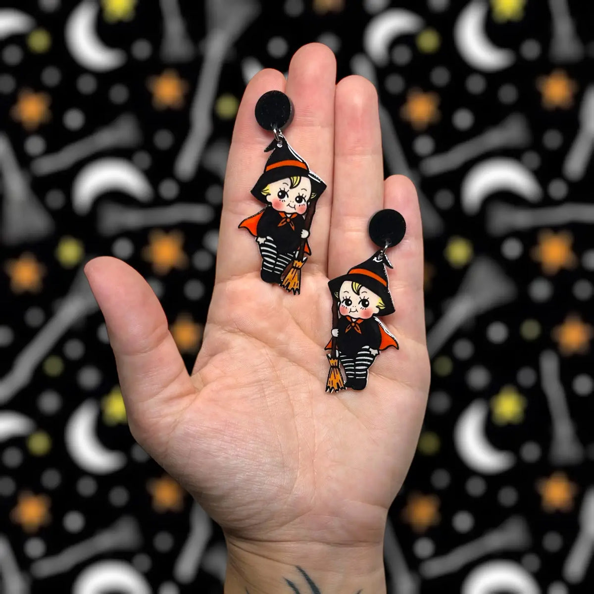 A hand holding two small witch-themed dangle earrings against a black patterned background with white crescent moons and yellow stars.