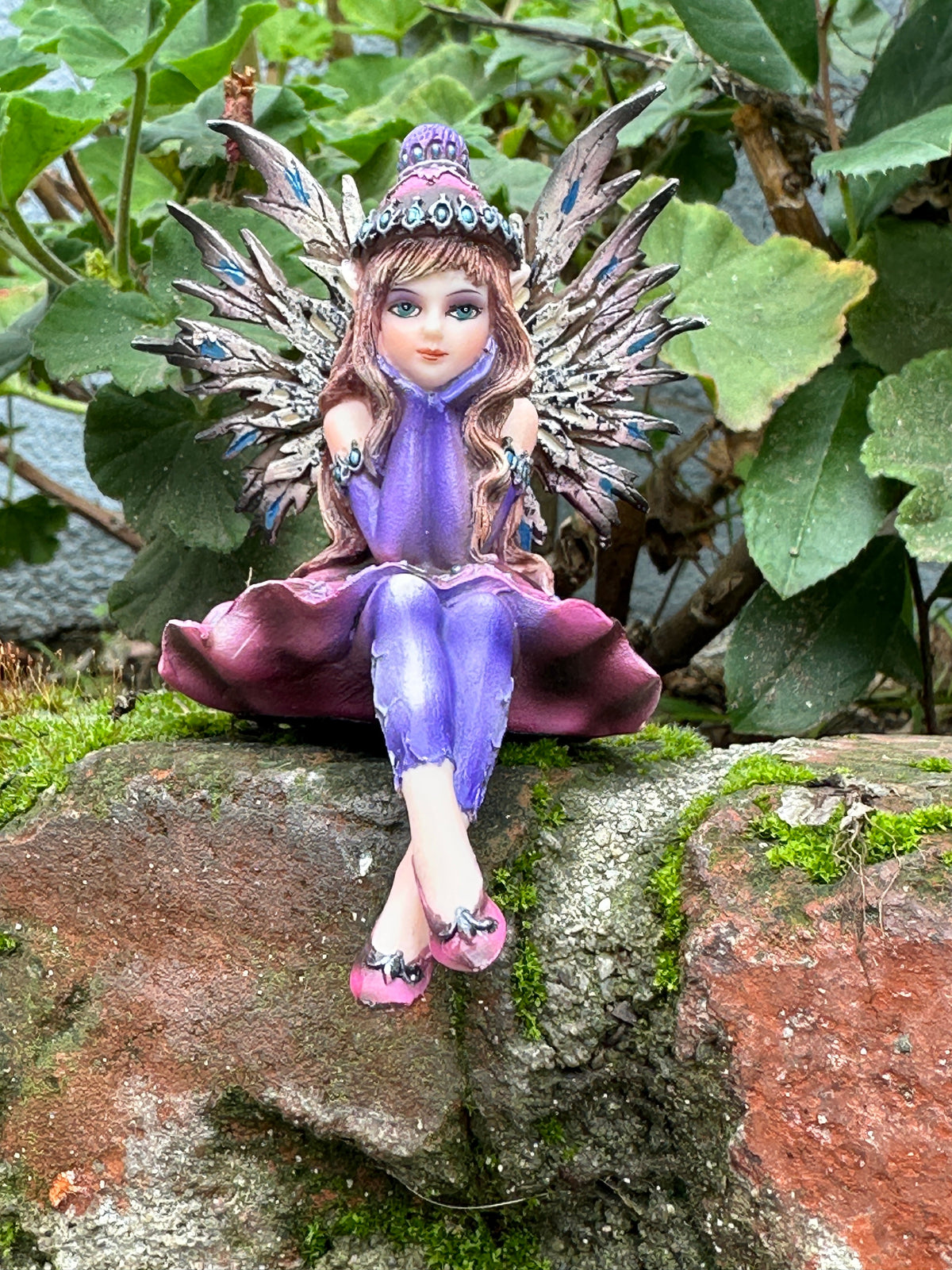 Amy Brown fairy figurine in purple dress and jeweled crown sitting on a mossy brick wall, surrounded by lush green leaves