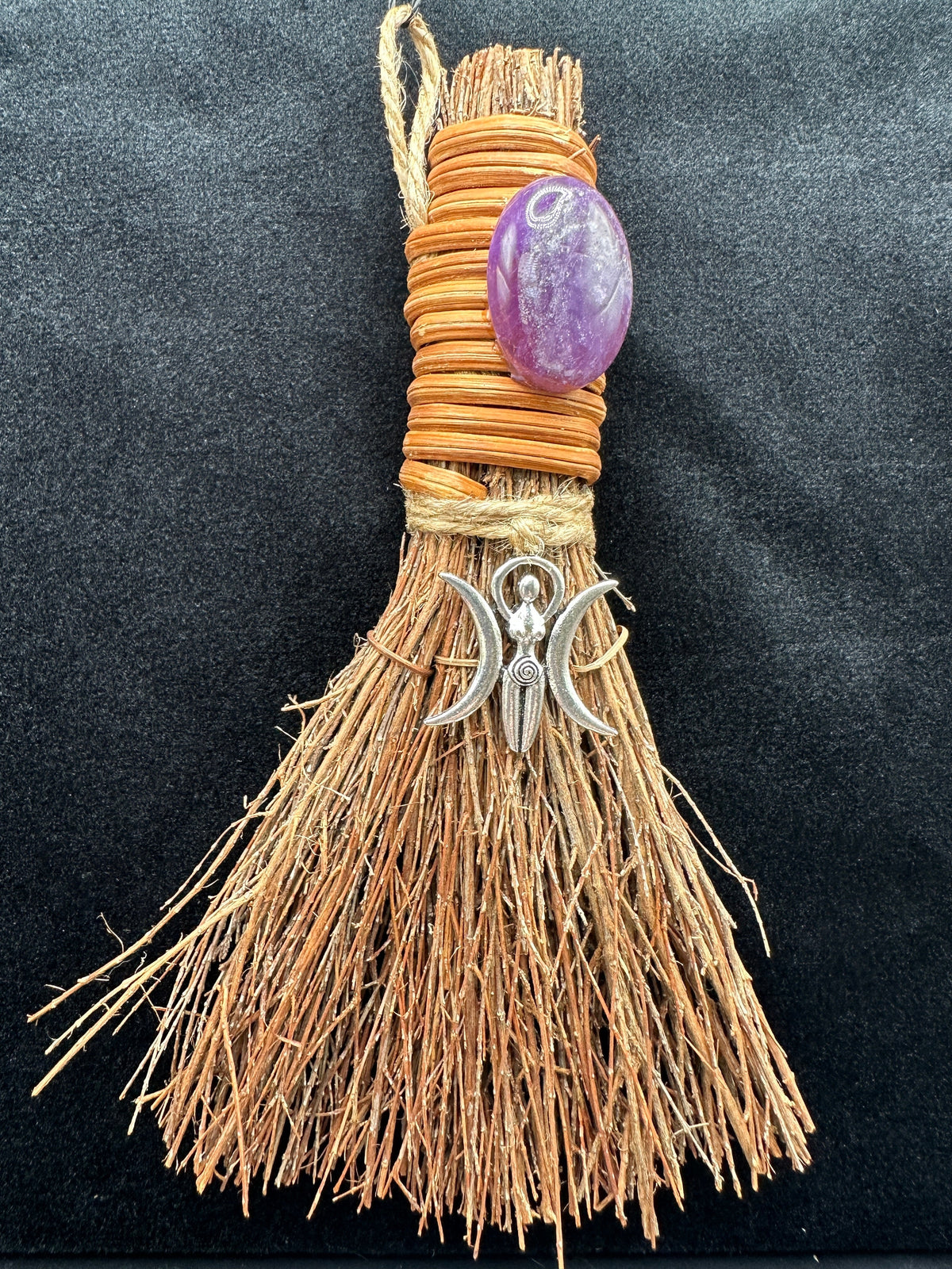 Handcrafted Triple Moon Goddess witch besom with natural raffia fibers, wrapped cord, polished amethyst crystal, and silver crescent moon charm on a dark background