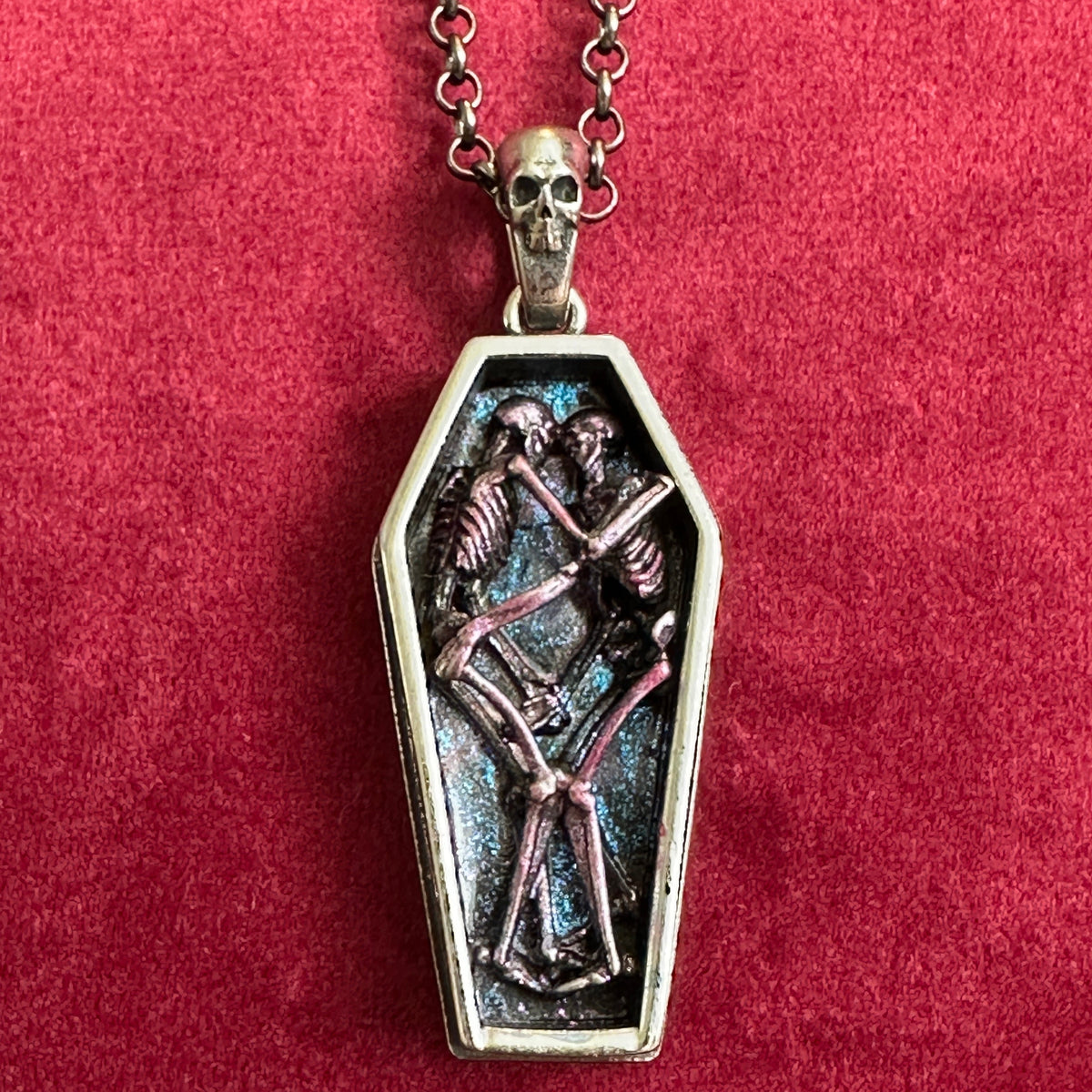 A gothic coffin pendant necklace featuring two interlocking purple skeleton figures on a silver chain featuring a silver skull displayed on a textured red velvet background.