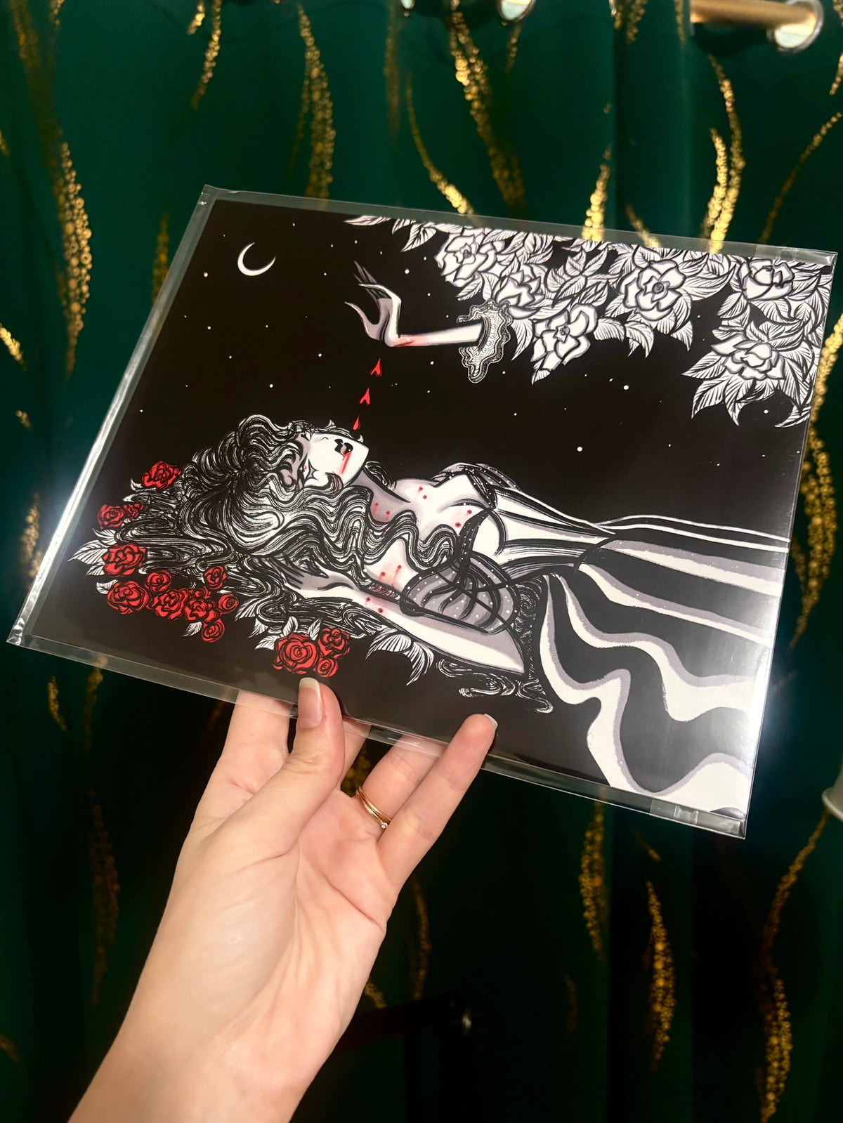 A hand holds a clear plastic sleeve containing a square art print or card featuring a black-and-white illustrated woman with red roses and a crescent moon, against a dark green curtain with gold vertical patterns.