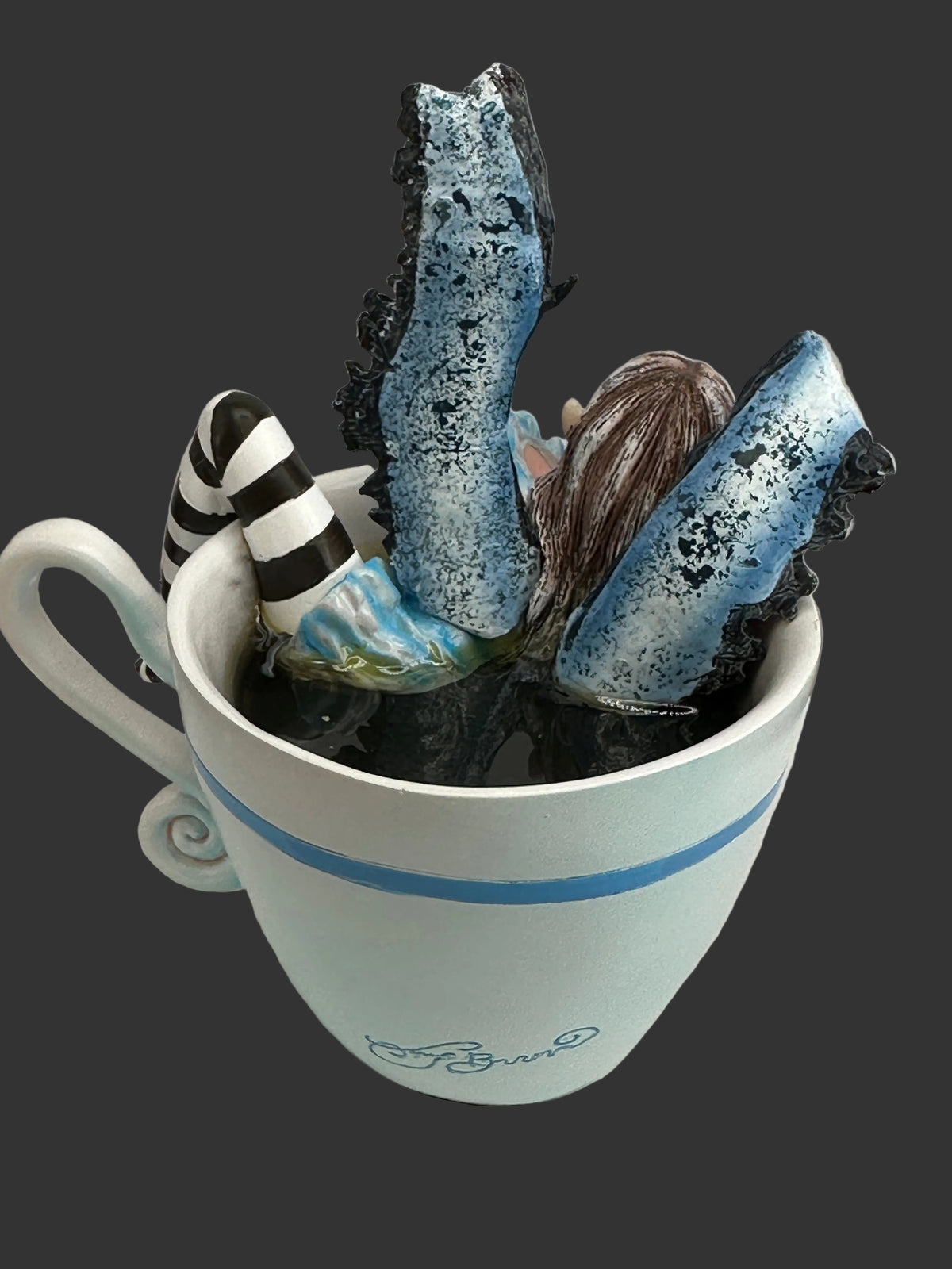 A ceramic mug with a blue stripe and the Amy Brown signature on the front, filled with whimsical decorative objects including blue-and-black textured pieces, a striped black-and-white item, and brown hair-like strands, set on a white surface against a light gray background.
