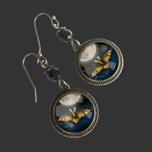 A pair of gothic-style dangle earrings featuring round bezel-set cabochons with a night sky scene of a bat flying in front of a full moon, suspended from antique bronze hooks with black faceted beads.