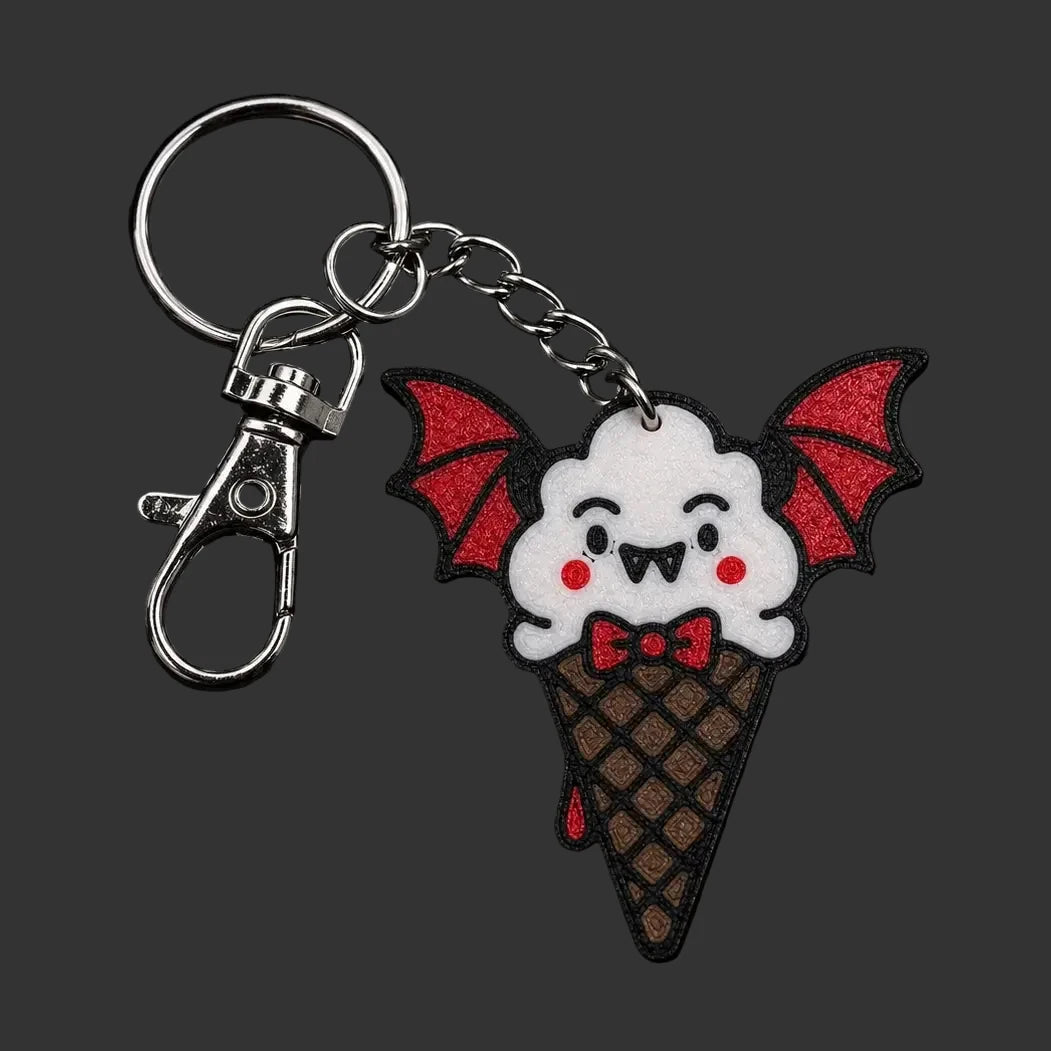 A cute bat-themed ice cream cone  gothic keychain charm  featuring a white bat face with red wings and a red bow tie on a brown waffle cone, attached to a silver metal clasp on a dark textured stone background.
