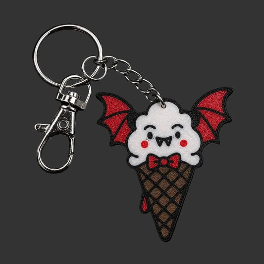 A cute bat-themed ice cream cone  gothic keychain charm  featuring a white bat face with red wings and a red bow tie on a brown waffle cone, attached to a silver metal clasp on a dark textured stone background.