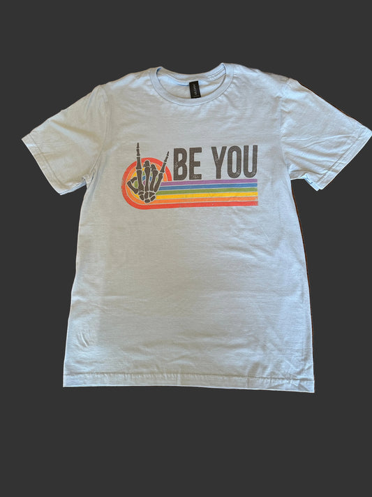 A light blue short-sleeve t-shirt laid flat on a wooden floor, featuring a graphic print with a skeleton hand giving the middle finger and the text 'BE YOU' above a rainbow stripe.