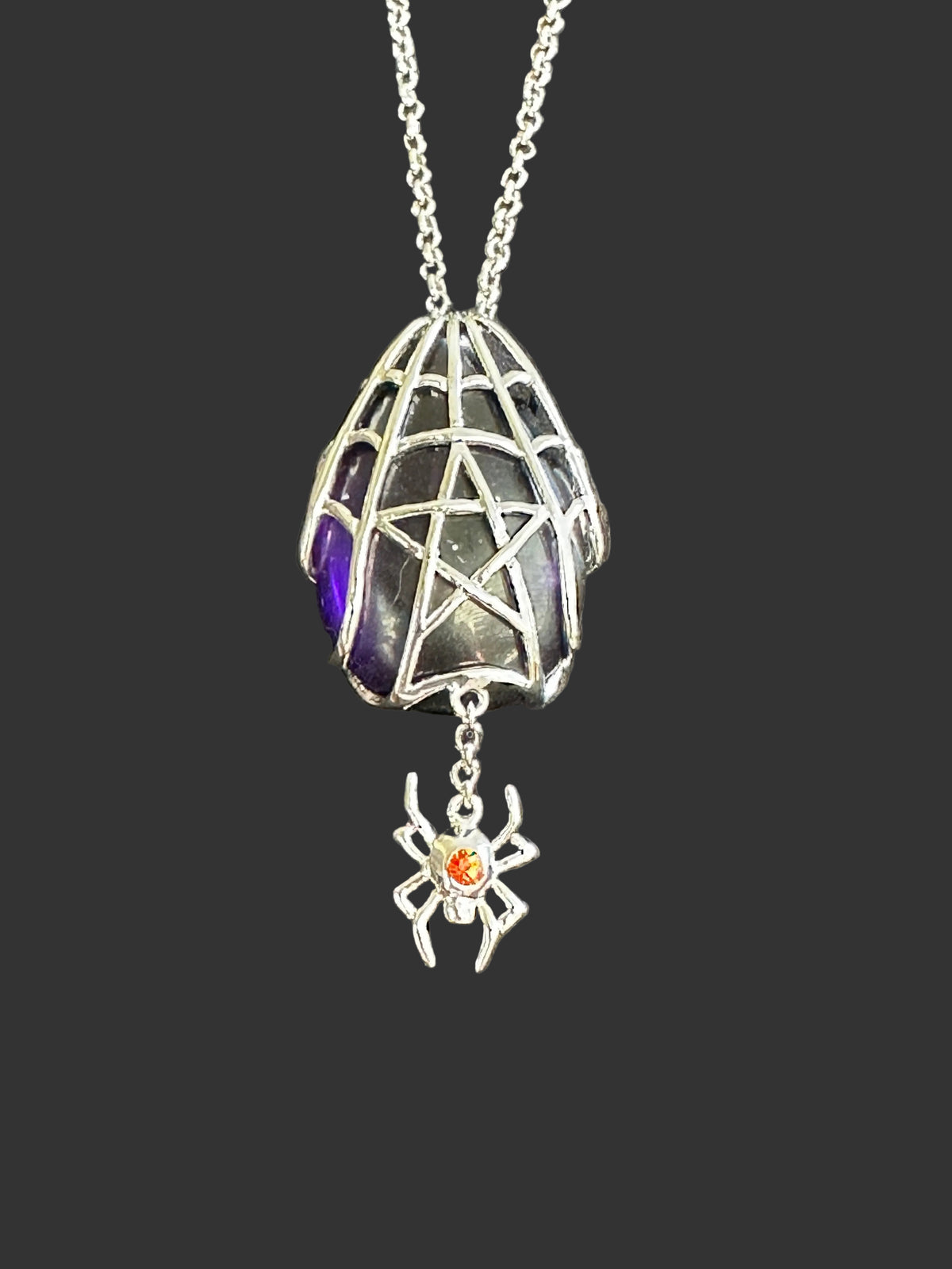An Anne Stokes gothic-style silver spider pendant necklace featuring a spiderweb cage design, a dark glass vial with purple and green iridescence, and a dangling silver spider charm with a small orange gemstone.