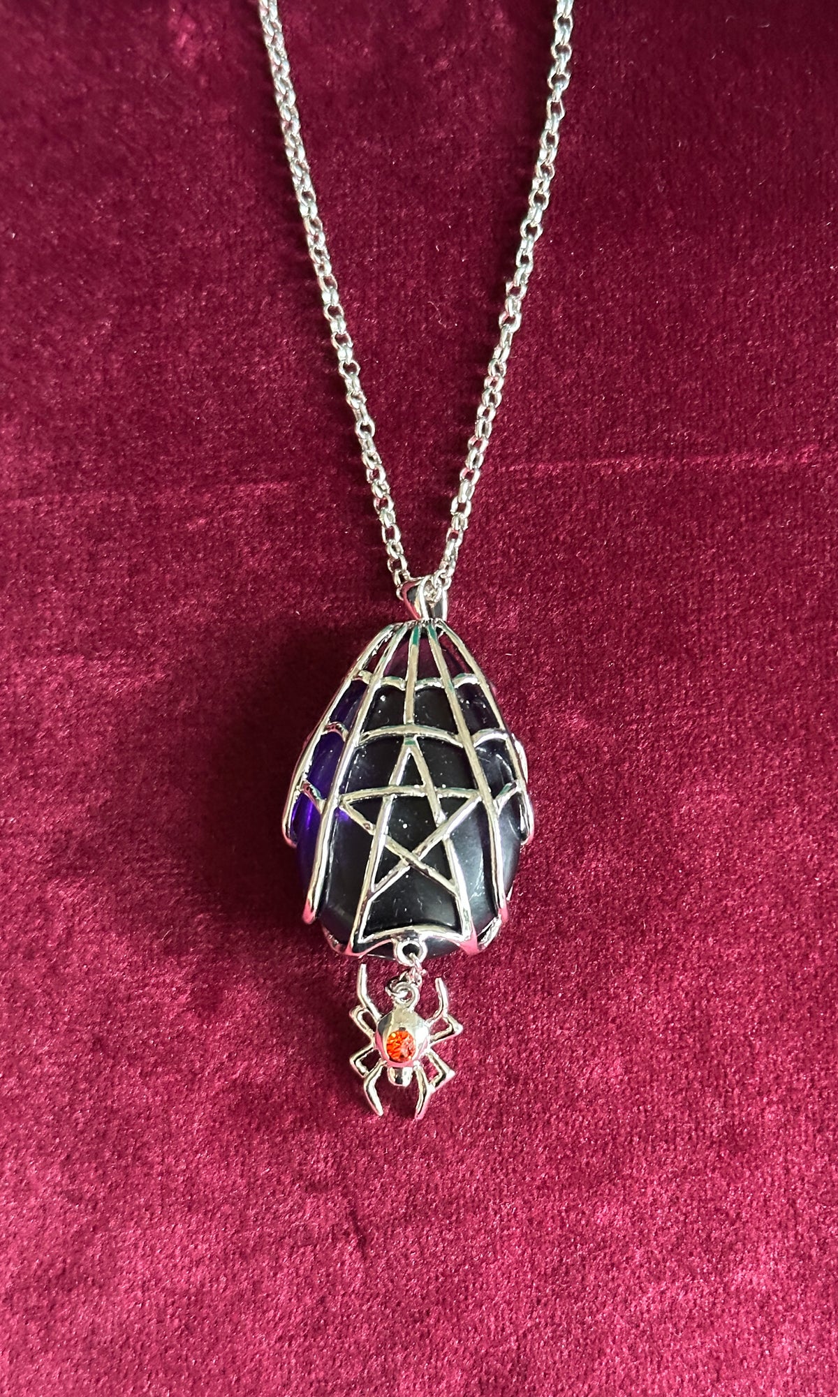 A silver-toned Anne Stokes pendant necklace featuring a spiderweb-patterned egg-shaped charm with a dangling spider accent holding a small orange gem, displayed on a deep burgundy velvet background.