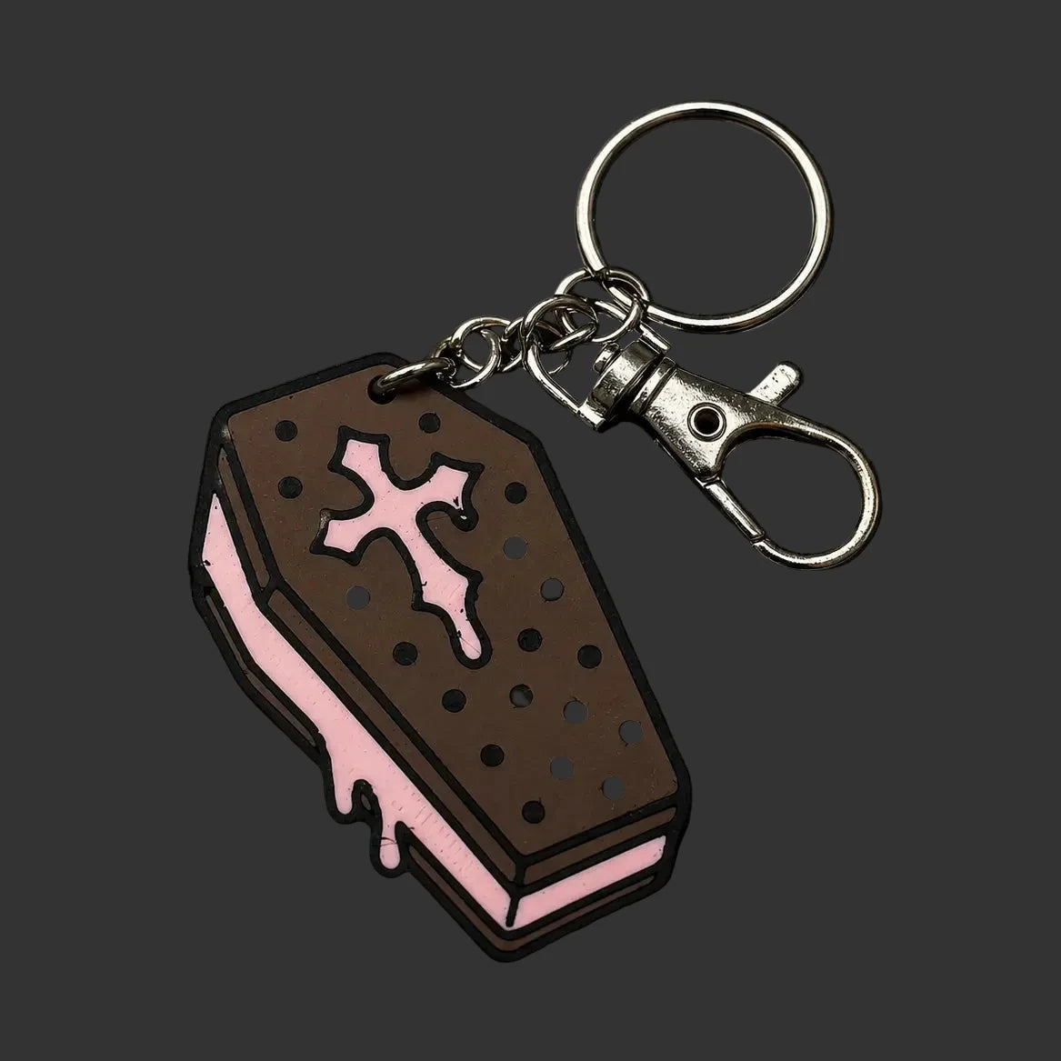 A coffin-shaped keychain charm styled like a chocolate sandwich cookie with pink icing and a pink cross, attached to a silver metal clasp and key ring, displayed on a dark textured slate surface.