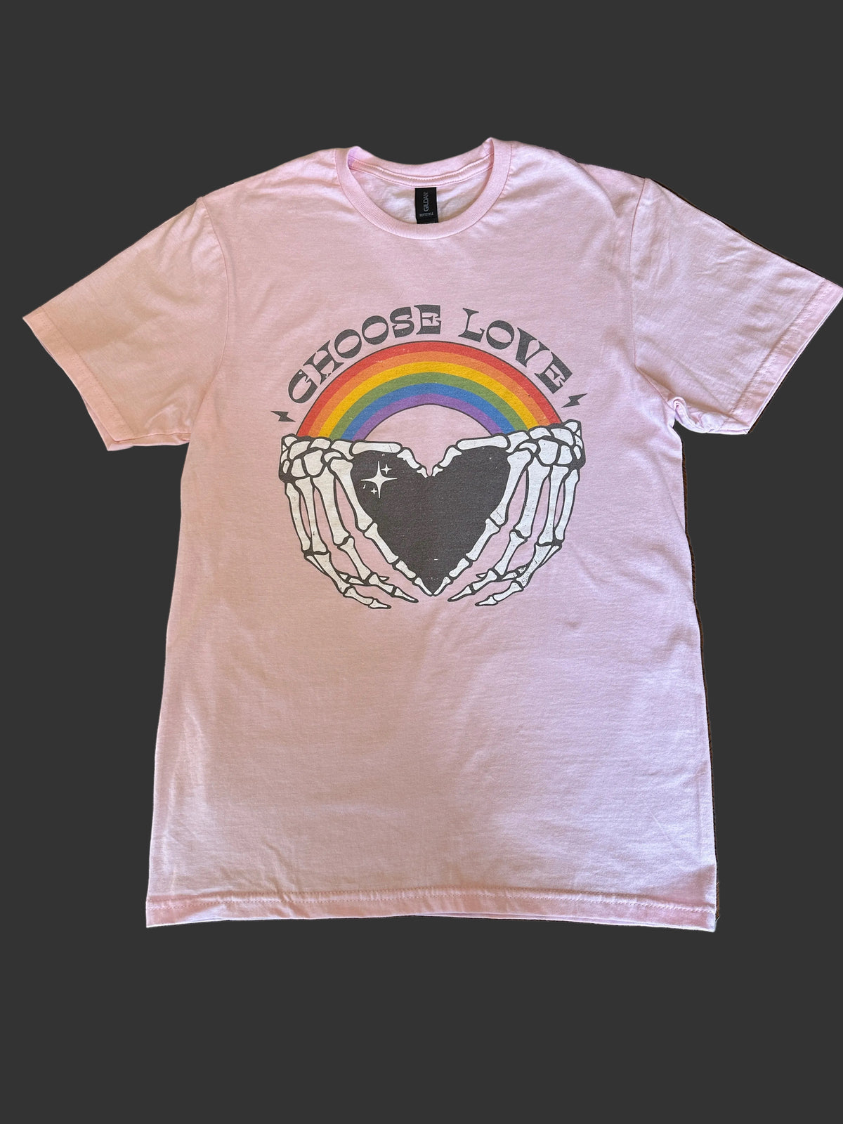 A light pink short-sleeve t-shirt laid flat on a wooden table, featuring a graphic of skeleton hands holding a black heart with a rainbow behind it and the words 'CHOOSE LOVE' arched above.