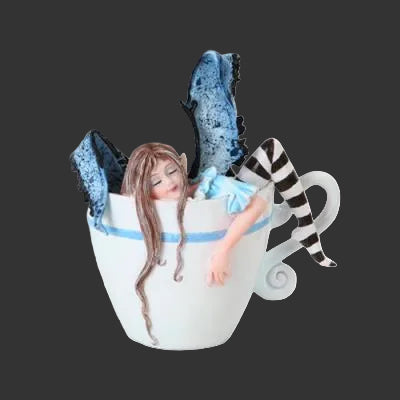 A whimsical Amy Brown figurine of a fairy with long brown hair, blue wings, and a black-and-white striped leg, nestled inside a large white teacup with a blue band.