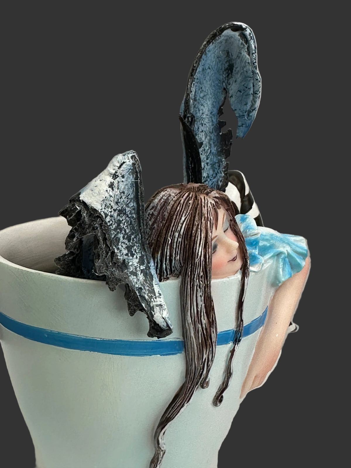 A ceramic fantasy figurine of a girl with long brown hair and blue wings, emerging from a white teacup with a blue stripe, set against a black background.