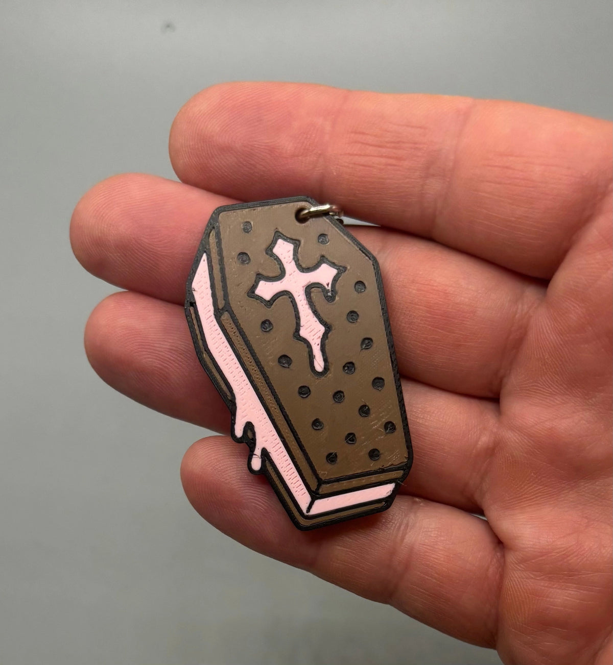 A hand holding a small coffin-shaped keychain or charm with a brown top, pink sides, and a white cross detail, shown against a plain gray background.