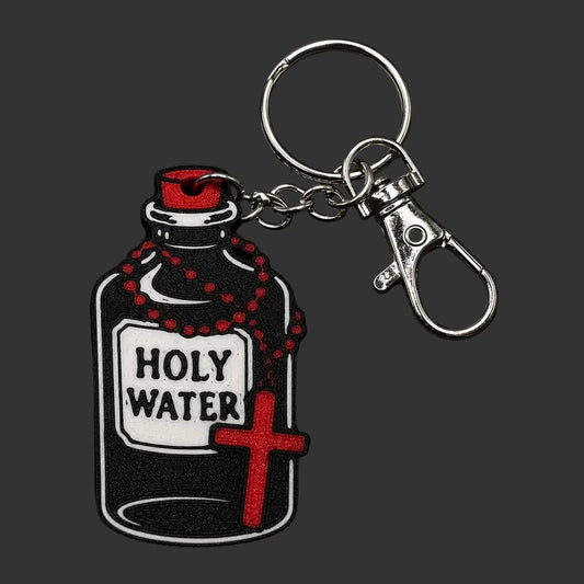 A Holy Water keychain featuring a glossy black and white bottle charm with a red cap, red rosary beads, and a red cross, attached to a silver metal split ring and lobster clasp, displayed on a dark textured slate background.