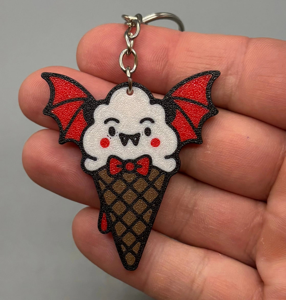 A hand holding a small bat-themed ice cream keychain. The keychain features a white cartoon ice cream scoop with red bat wings, a red bow tie, and a brown waffle cone, attached to a silver metal key ring.
