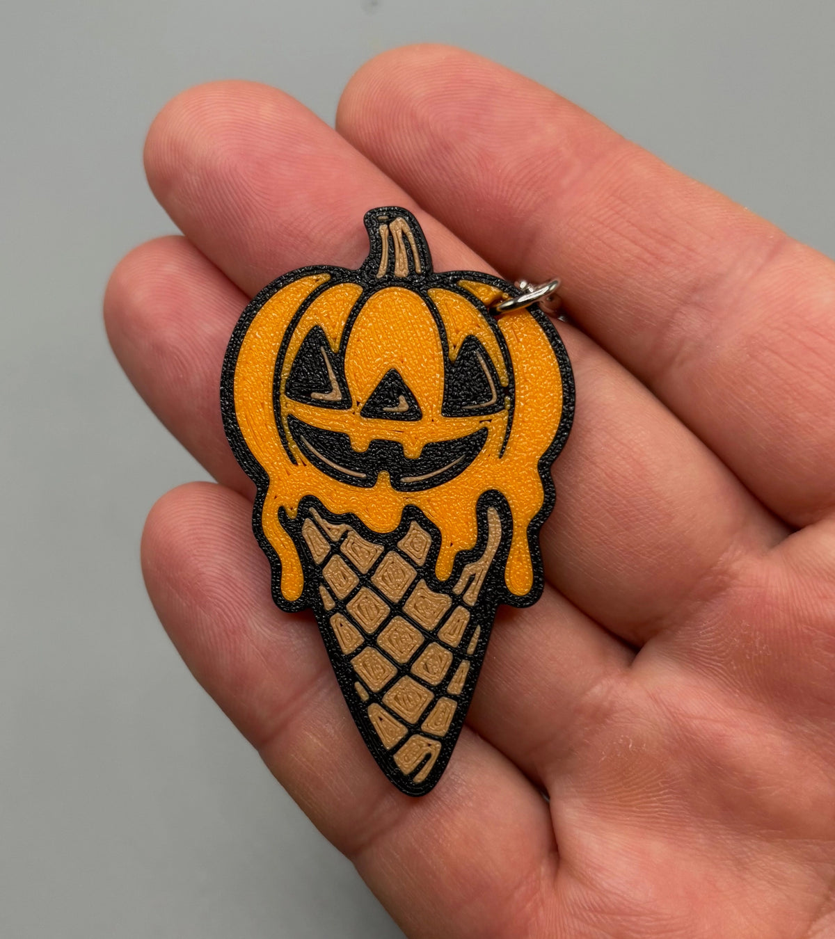 A Halloween-themed charm or keychain shaped like a jack-o'-lantern pumpkin dripping into an ice cream cone, held in a person's hand against a plain gray background.