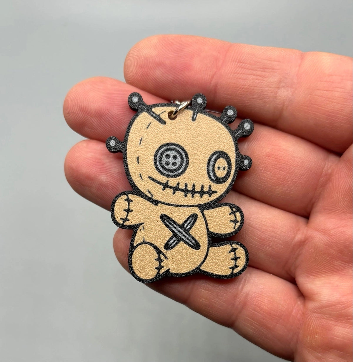 A small voodoo doll keychain or charm held in a person's hand. The charm is tan with black stitched details, a button eye, and a pinhead design, with a silver metal ring at the top.