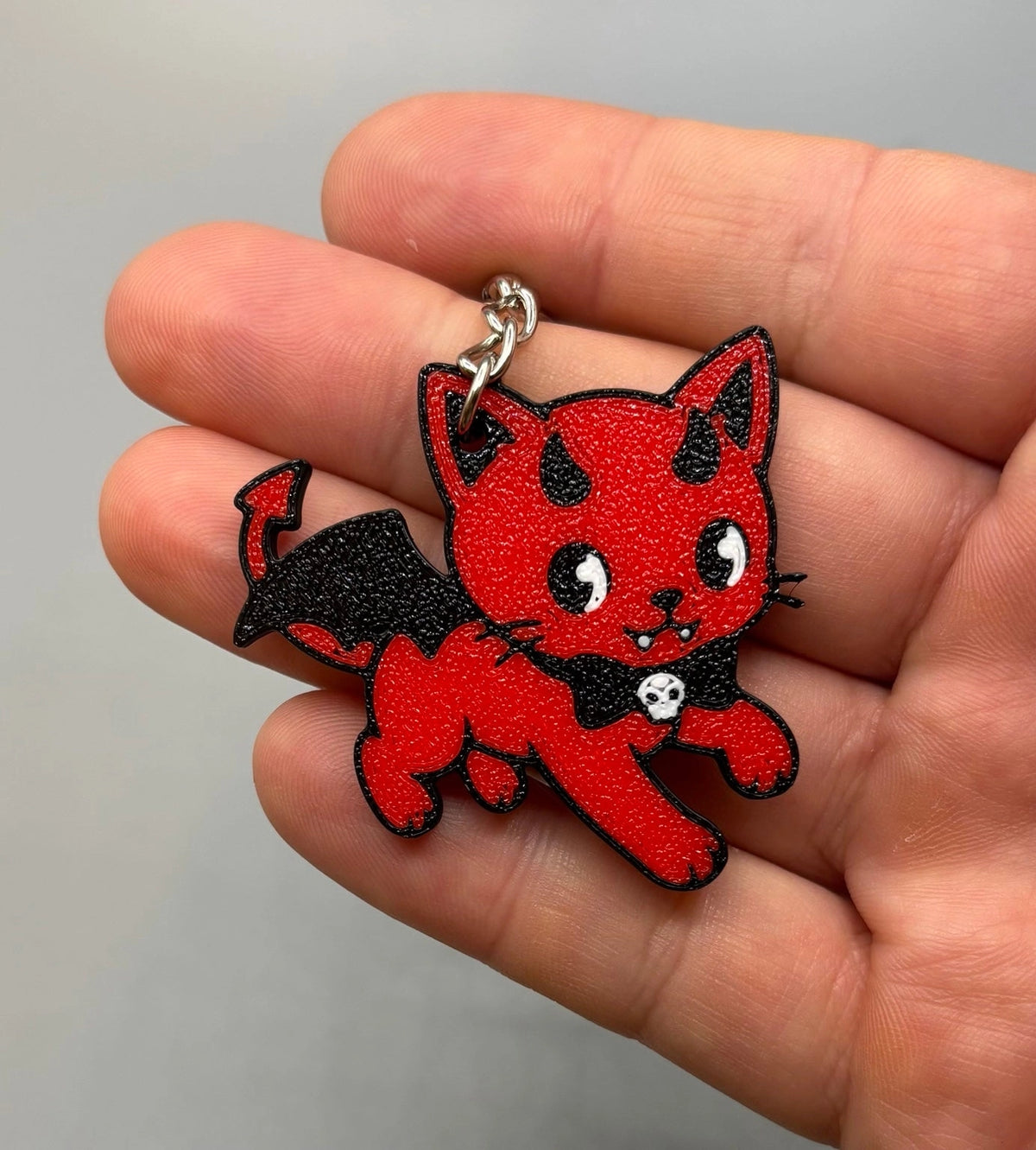 A close-up of a hand holding a small red devil kewpie cat keychain charm with black wings, black outlines, and a silver metal ring.