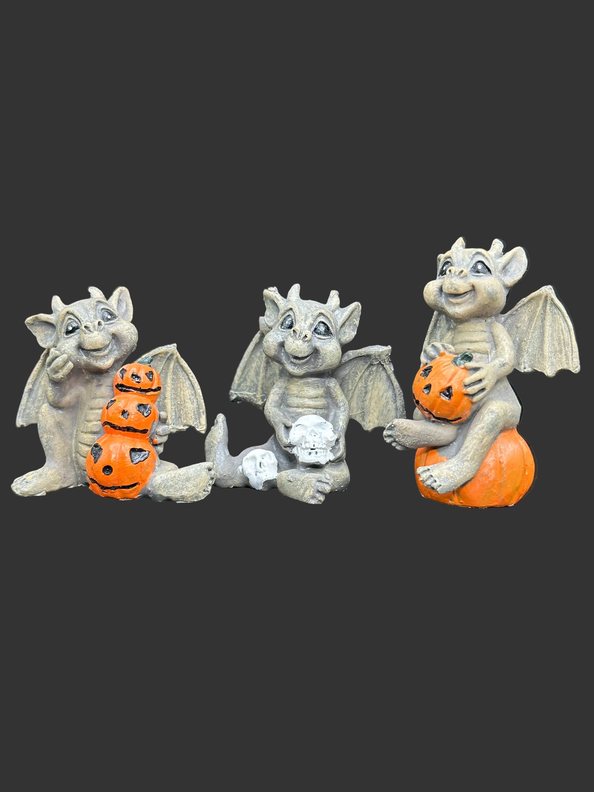 Three small gargoyle figurines in a weathered gray finish, each holding orange jack-o'-lantern pumpkins, displayed on a dark reflective surface against a dark gray background.
