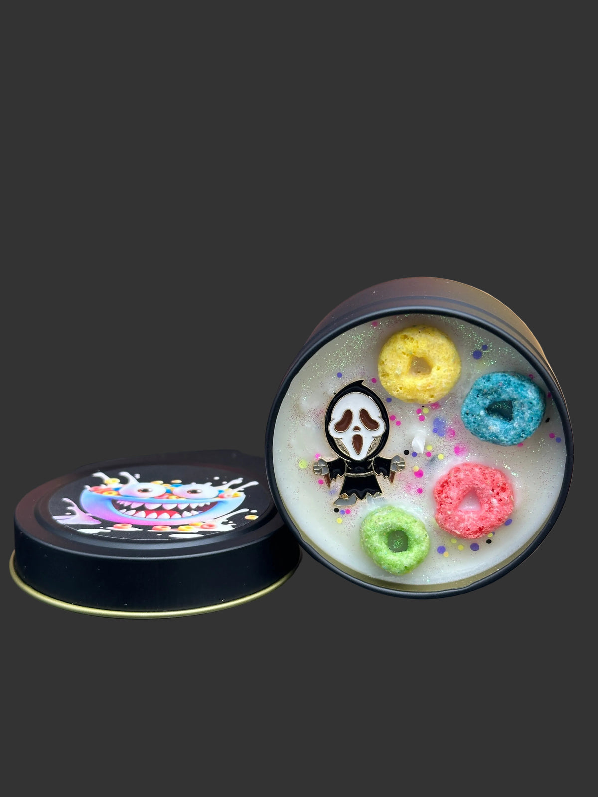 A round black tin container shown open, revealing a white wax melt or soap insert with colorful cereal-ring shapes and a ghost-face style character. The matching black lid sits beside it on a dark surface, with a blurred Halloween-themed background featuring pumpkins and a house.