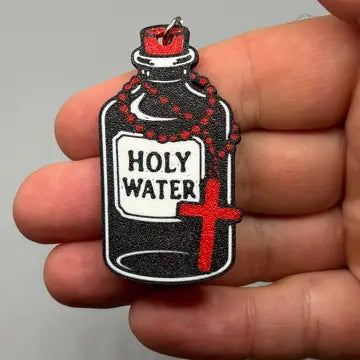 A small 3D printed charm or keychain shaped like a bottle labeled 'HOLY WATER' with a red cross and rosary, held in a person's hand against a plain light gray background.