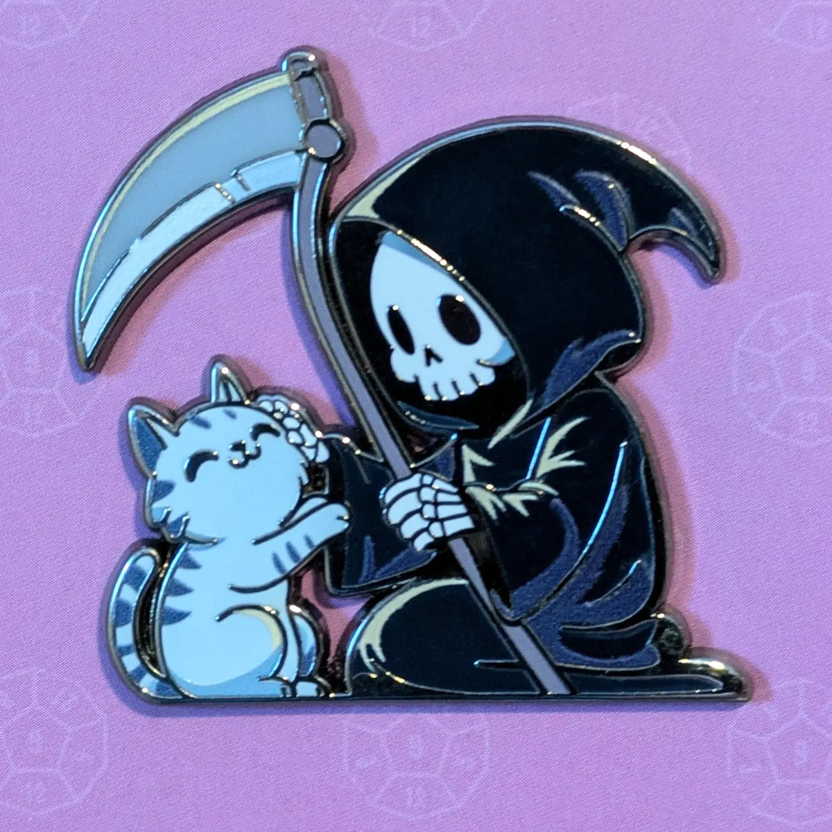A cartoon grim reaper and cat enamel pin on a purple patterned background. The pin features a black hooded reaper holding a silver scythe next to a light white and grey striped cat.