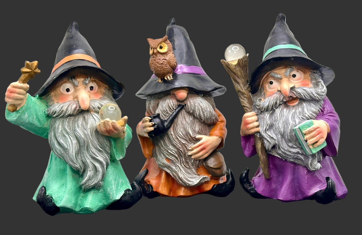 Three whimsical wizard garden gnomes with long gray beards and black pointed hats, standing on a black surface against a white background. The left gnome wears a green robe and holds a star and a crystal ball; the center gnome wears an orange robe with a purple hat and an owl on its hat; the right gnome wears a purple robe with a green hat and holds a staff and a book.