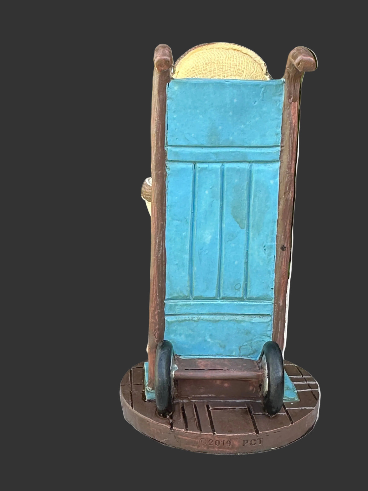 A small cold cast resin collectible Pinhead Monster of a Hannibal Lector on a blue rolling door with brown wooden side rails and wheels, sitting on a weathered wooden table with green foliage in the background.