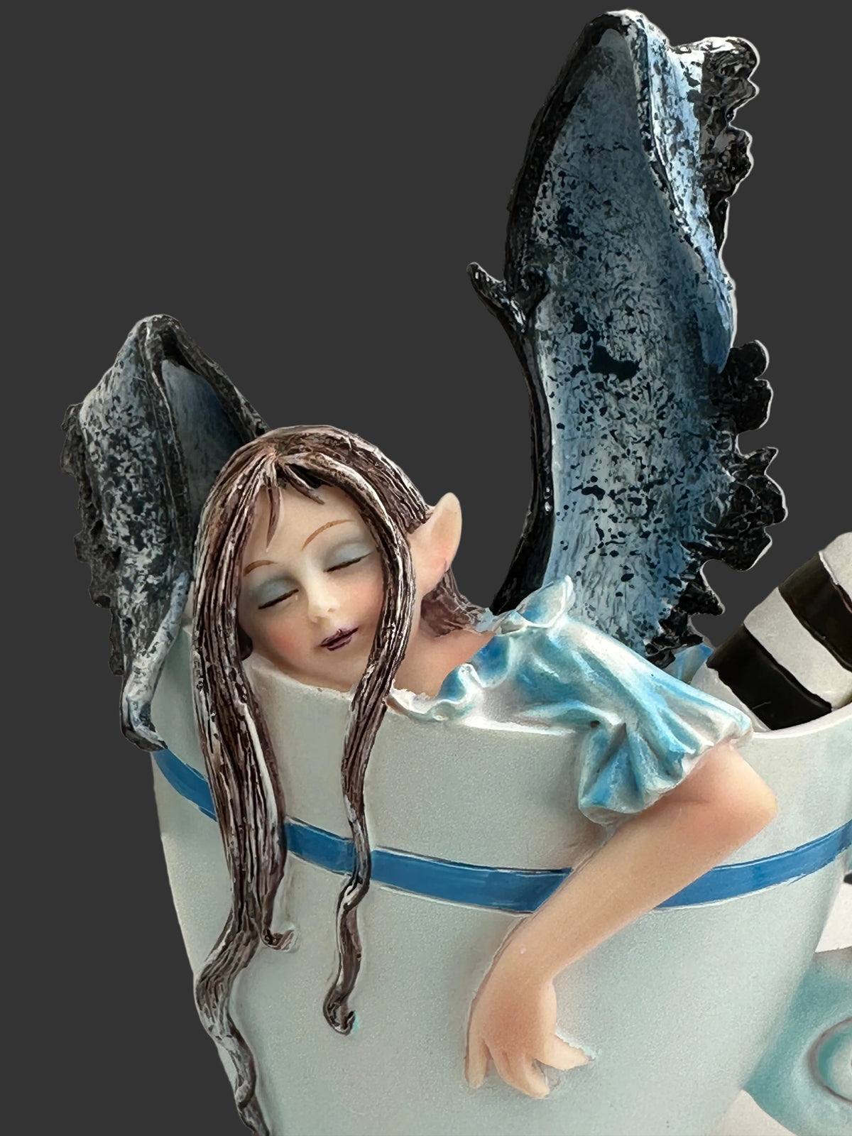 A fantasy Amy Brown figurine of a sleeping female fairy with long brown hair, pointed ears, and blue wings, emerging from a large white teacup with a blue stripe.