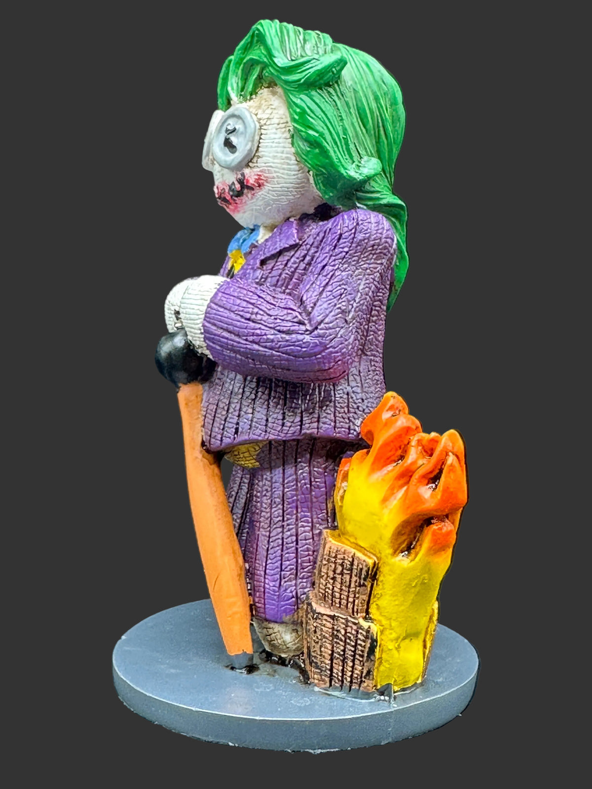 A painted collectible figurine of the Joker from Batman, holding a orange cane, standing next to a yellow and orange pile of ashes on a gray circular base against a black background.