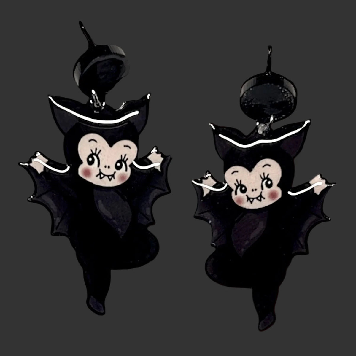 Two black bat-shaped hanging black kewpie vampire babies with white trim and cartoon faces, shown against a dark moody black background