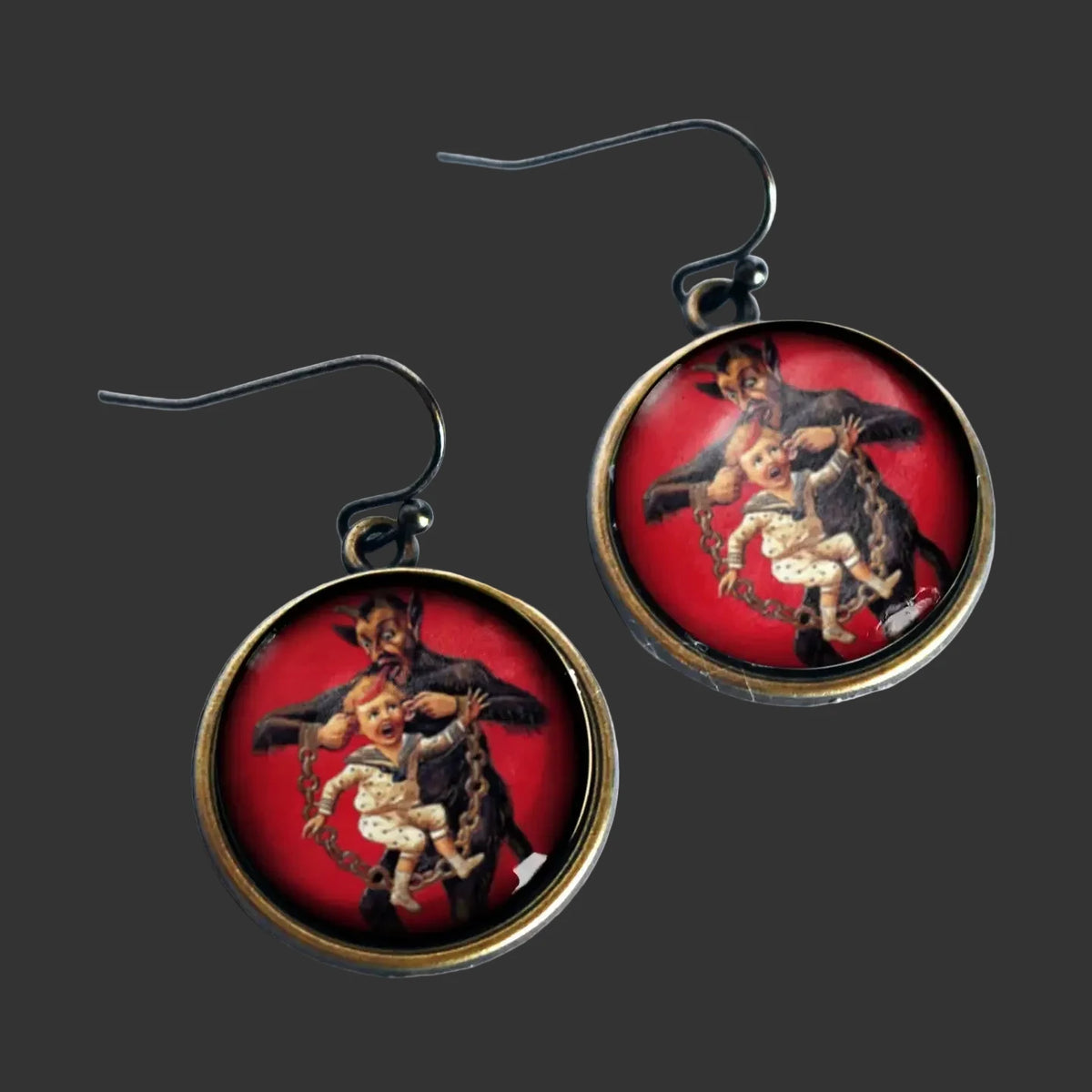 A pair of round drop earrings with black fishhook wires and bronze-toned bezels, featuring a vintage-style illustration of a Krampus, a dark-haired figure holding a child against a red background, shown on a black background.
