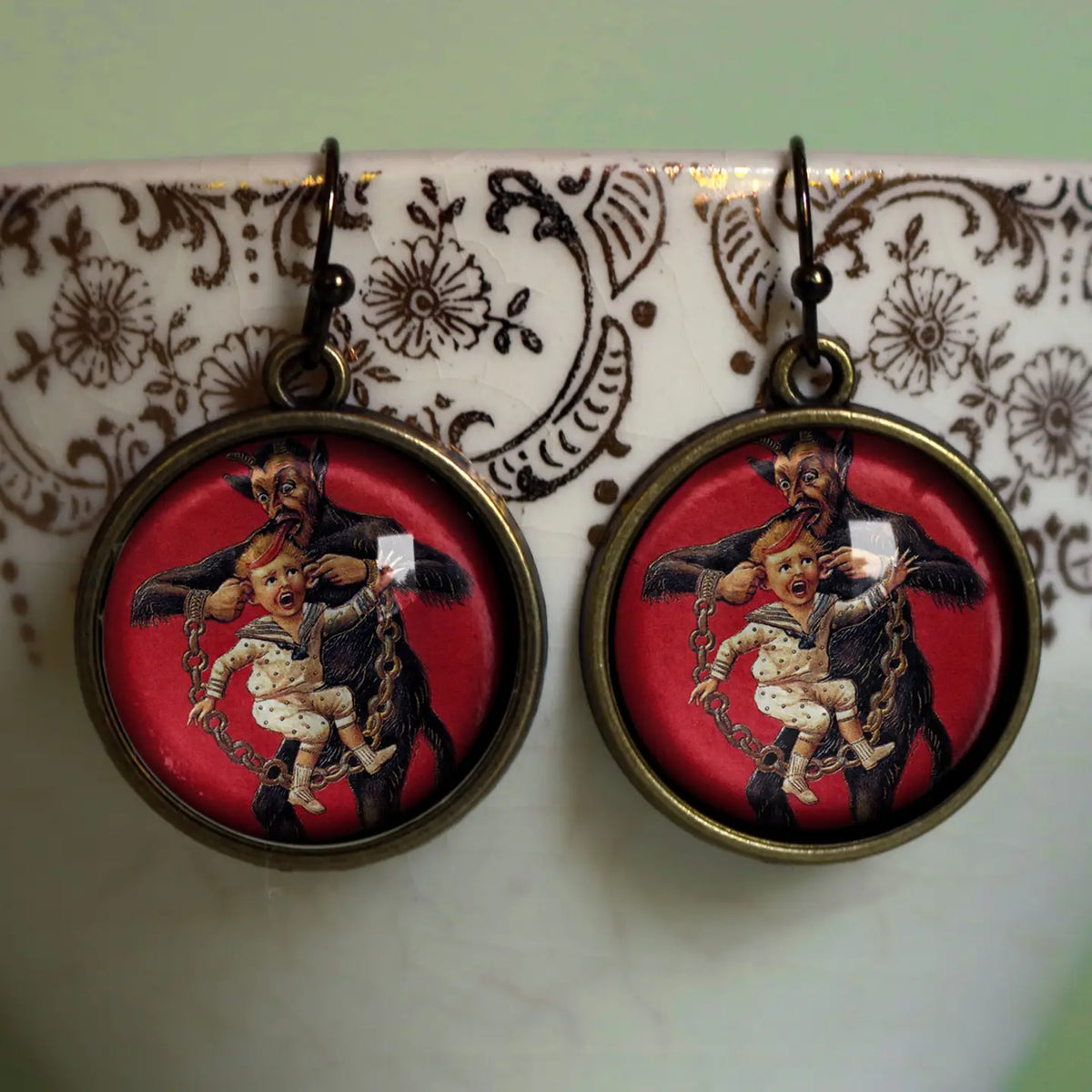 A pair of round drop Christmas earrings with antique bronze-toned metal frames and hooks, featuring a vintage-style illustration of Kramous,  dark-haired man holding a child against a red background, displayed on a white ceramic surface with a brown floral pattern.