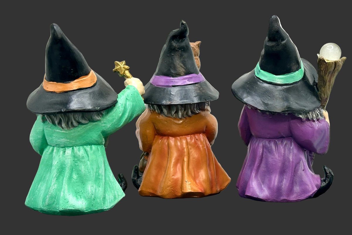 Three small warlock miniatures shown from the back, each wearing a pointed black hat and long dress in green, orange, and purple, standing on a dark round surface against a light gray background.