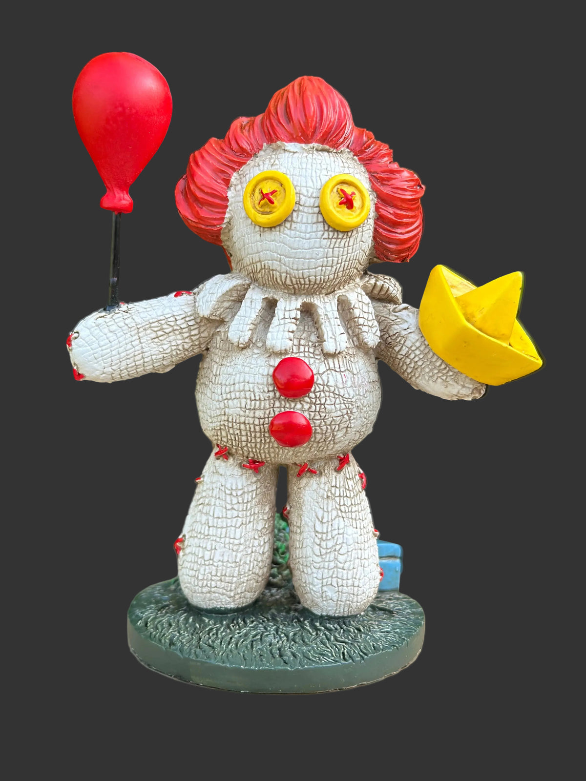 A creepy clown figurine standing on a textured green base, holding a red balloon and a yellow paper boat. The clown has a textured off-white body, bright red hair, yellow starry eyes, and red buttons, set against a blurred green foliage background.