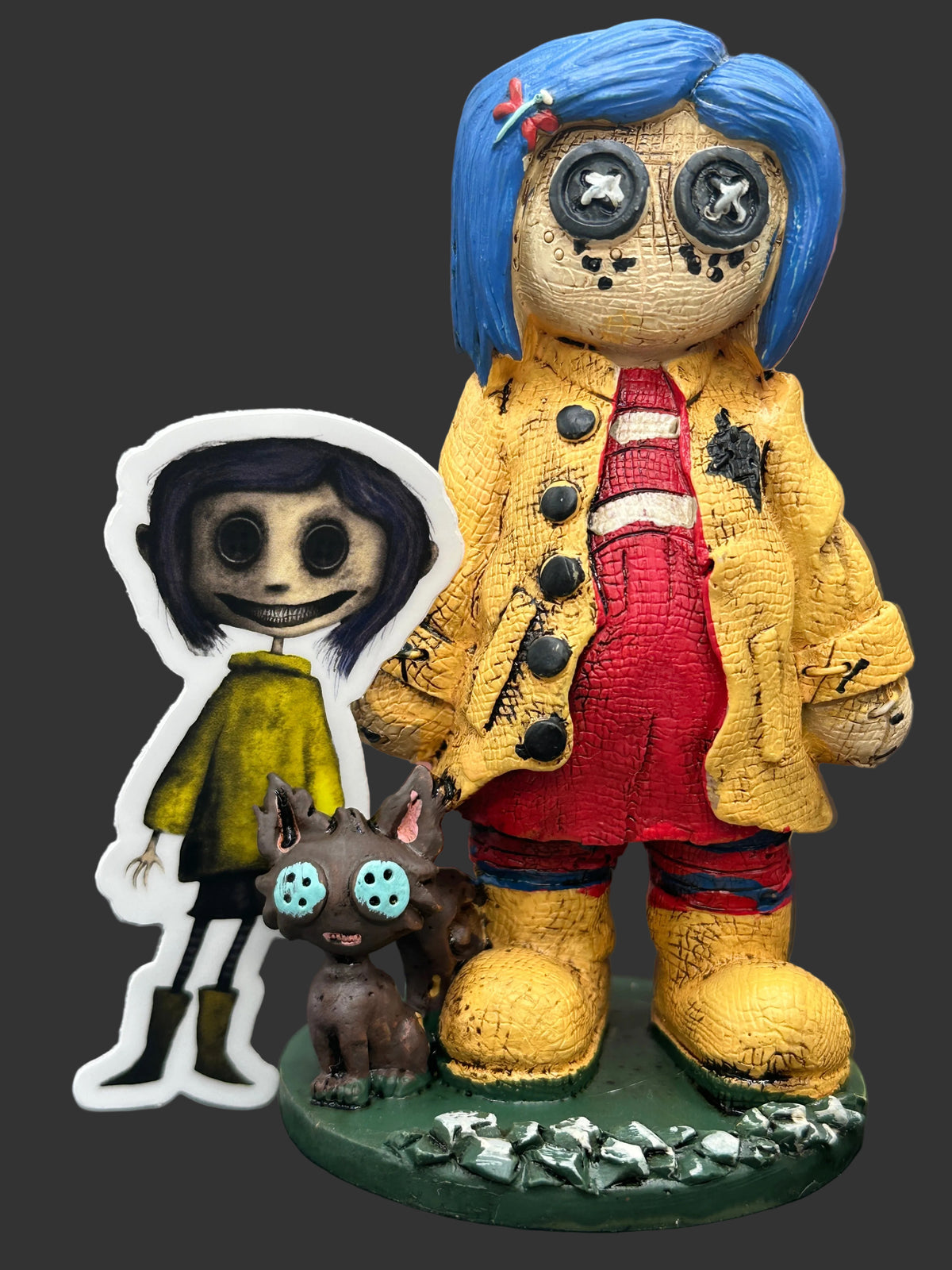 A horror-themed collectible Pinhead Monster Coraline figurine set featuring a creepy blue-haired doll in a yellow coat and red overalls on a green base, alongside a die-cut Creeptoons Coraline sticker and a small brown cat figure with blue eyes.
