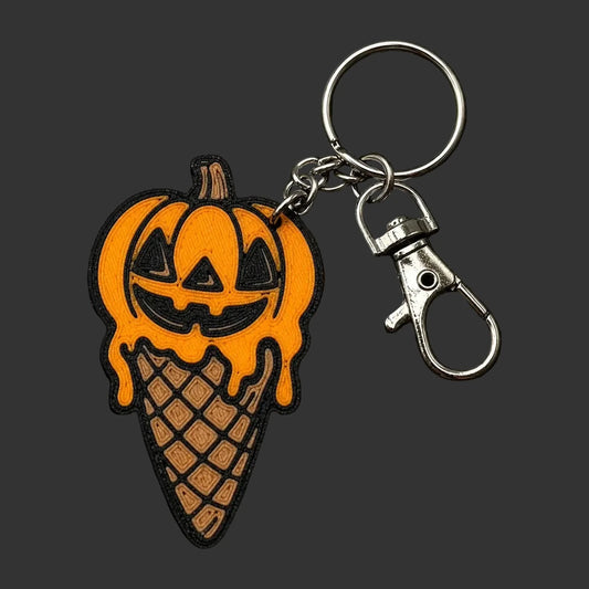 A Halloween-themed keychain featuring a jack-o'-lantern pumpkin dripping into an ice cream cone, attached to a silver metal split ring and lobster clasp, displayed on a dark textured stone coaster.